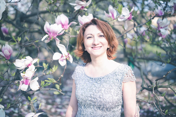 Radosna Ormianka w średnim wieku w eleganckiej sukience pod kwitnącym drzewem magnolii. - Zdjęcie, obraz