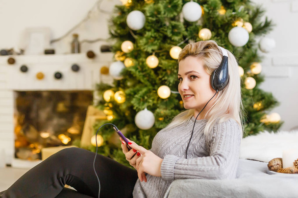 Donna felice godendo la sua musica a Natale rilassante su un divano di fronte all'albero con un sorriso raggiante stringendo il suo cellulare al petto
. - Foto, immagini