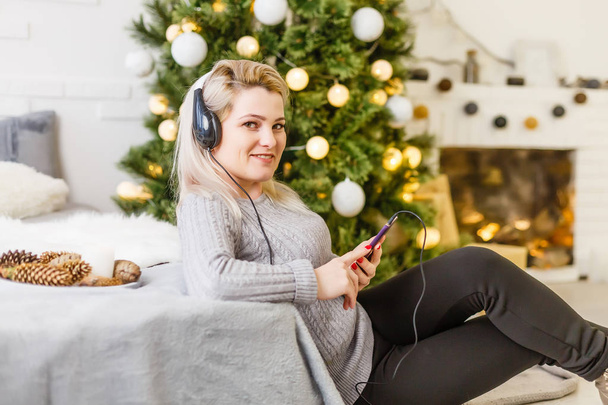 Щаслива жінка насолоджується своєю музикою на Різдво, розслабляючись на дивані перед деревом з блимаючою посмішкою, що прив'язує її мобільний до грудей
. - Фото, зображення