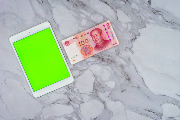Finanzas Concepto de negocio. Un billete de banco chino Yuan RMB y una tableta de pantalla táctil de pantalla verde en la mesa
. - Foto, imagen