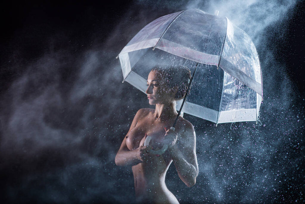 A beautiful sad nude girl takes shelter from a shower at night under a transparent umbrella. Lonely woman in the pouring rain in the light of spotlights and flashes. Water drops. - Foto, Imagem