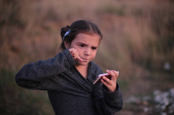Egy hét éves lány, különböző érzelmekkel az arcán játszik a telefonban. - Fotó, kép