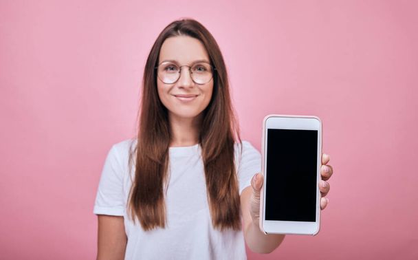 Cool joyful girl in a white t-shirt standing and shows phone display right in the frame - Foto, afbeelding