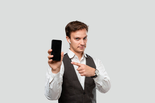 Όμορφος επιχειρηματίας με λευκό πουκάμισο και γκρι γιλέκο που δείχνει την οθόνη ενός smartphone. - Φωτογραφία, εικόνα