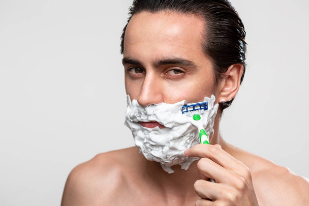 Bıyıklı yakışıklı bir adam beyaz arka planda yalın ayak duran sakalını kesmek için sopa ve tıraş köpüğü kullanır. Sabah tedavisi ve tıraş kavramı. - Fotoğraf, Görsel