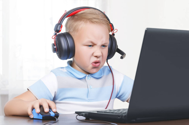 Pequeño chico lindo europeo hermoso se sienta en un ordenador portátil en los auriculares con un micrófono con un ratón de ordenador en la mano y juega un juego en línea. El niño está enojado e insatisfecho con su pérdida y cl
 - Foto, imagen