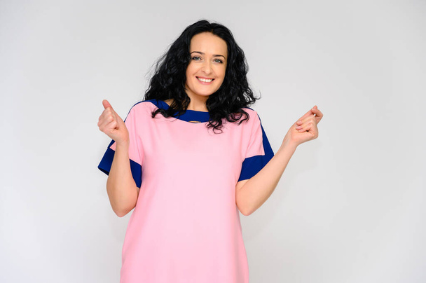 Portrait of a pretty brunette woman with long curly hair on a white background in a pink dress. He stands in front of the camera, smiles, talks in various poses. - Fotoğraf, Görsel