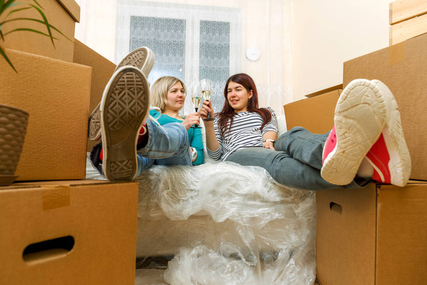 Foto von zwei Mädchen mit Weingläsern sitzt auf Sofa zwischen Pappkartons in neuer Wohnung - Foto, Bild