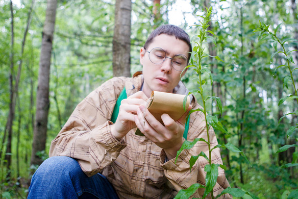 Photo of dark-haired biologist in glasses writes in notebook in forest among plants and trees in summer afternoon - Fotó, kép