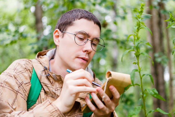 Image d'un biologiste aux cheveux bruns avec des lunettes écrivant avec un crayon dans un cahier en forêt parmi les plantes et les arbres l'après-midi d'été
 - Photo, image