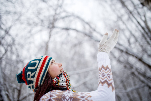 Fénykép mosolygós lány kötött kalap és sál fogása hópelyhek téli erdőben a nap folyamán, disokted háttér - Fotó, kép
