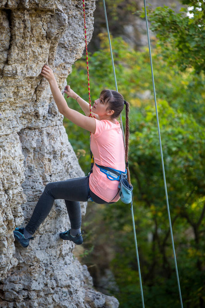 Foto da menina atleta que clambering sobre a rocha contra o fundo de árvores verdes
 - Foto, Imagem