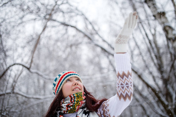 Photo of smiling brunette looking up in knitted hat and scarf catching snowflakes in winter forest during day, defocused background - Photo, Image