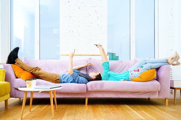 Ο τύπος και το κορίτσι ξαπλωμένος στον καναπέ, συζητώντας κάτι στο smartphone. Έννοια των κοινωνικών μέσων μαζικής ενημέρωσης. - Φωτογραφία, εικόνα