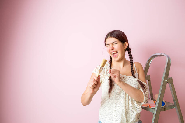 Фото веселої жінки з пензлем поруч зі драбиною і валиком проти порожньої рожевої стіни. Місце для тексту
. - Фото, зображення
