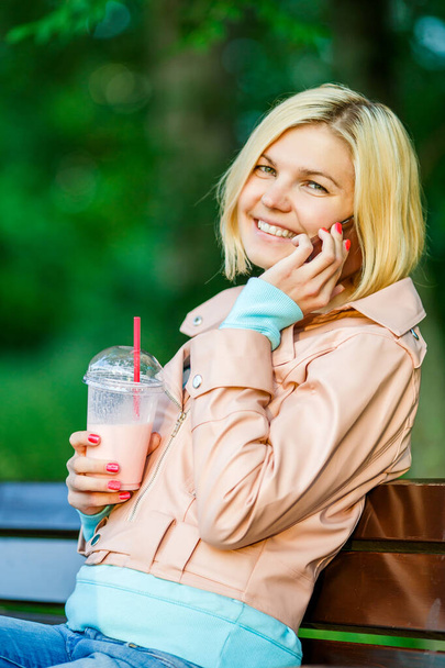 Blonde with milkshake talking on phone while sitting at bench, blurred background - Photo, Image
