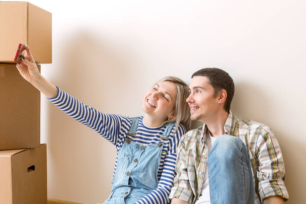 Φωτογραφία του νεαρού παντρεμένου ζευγαριού να κάνει selfie κάθεται στο πάτωμα ανάμεσα σε κουτιά από χαρτόνι στο νέο διαμέρισμα - Φωτογραφία, εικόνα