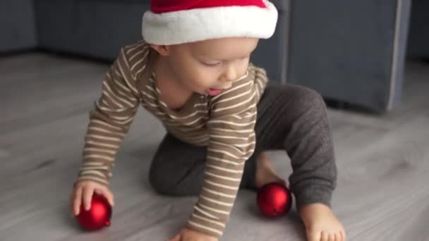 Toddler in a Santas fleece hat is playing with a red Christmas ball. Merry Christmas and Happy New Year - Materiał filmowy, wideo