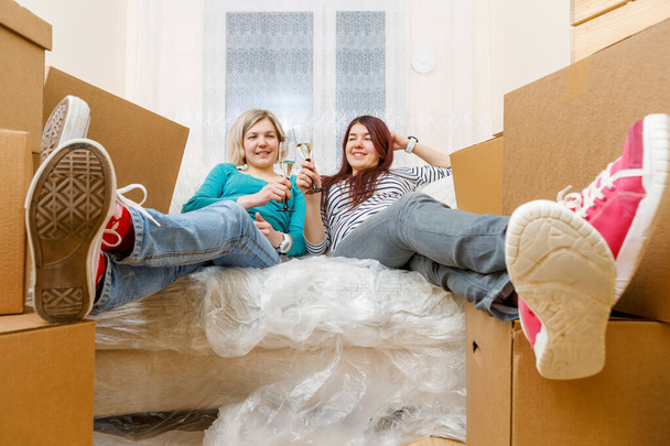 Photo of two girls with glasses of wine sitting on sofa among cardboard boxes in new apartment - Valokuva, kuva