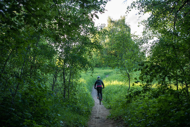 Фото ззаду жінки велосипедист в шоломі їде по дорозі в лісі вдень
 - Фото, зображення