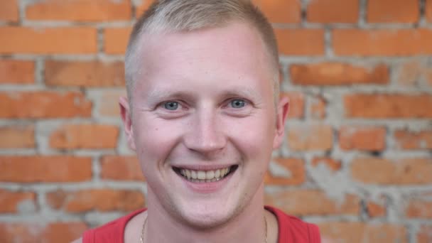 Close up of happy young man looking into camera with positive emotions on his face outdoor. Portrait of smiles strong sportsman with blonde hair standing on blurred brick wall background - Footage, Video