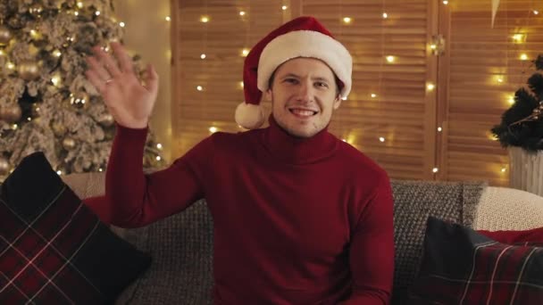 Portrait of Young Cheerful Man Wearing Santas Hat and Winter Sweater Sitting on the Sofa Holding Present and Waving Hi with Christmas Tree at the Background - Video