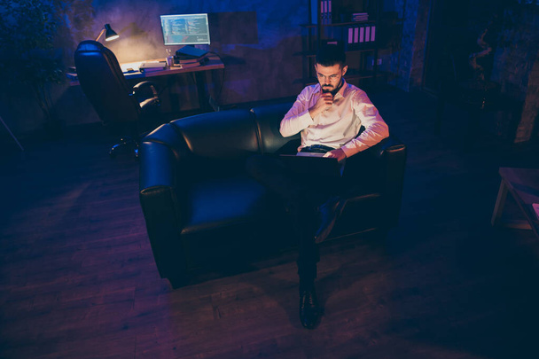 Teljes hossza keményen dolgozó üzletember ül a bőr kanapén munka laptop olvasott projekt alkalmazottai úgy vélik, elemezni információkat marad este sötét iroda padláson - Fotó, kép