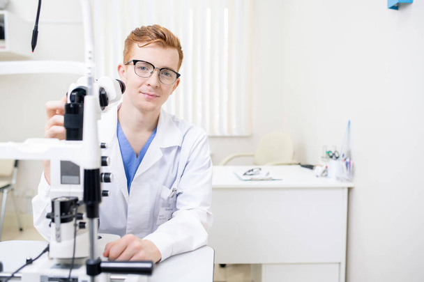 Νεαρός επιτυχημένος οφθαλμίατρος με στολή κάθεται στο χώρο εργασίας ενώ δοκιμάζει νέο οφθαλμολογικό εξοπλισμό στην εργασία - Φωτογραφία, εικόνα