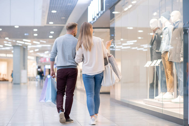 Achteraanzicht van jonge shoppers die aandacht besteden aan nieuwe mannelijke casualwear collectie in etalage terwijl ze tijd doorbrengen in het winkelcentrum - Foto, afbeelding