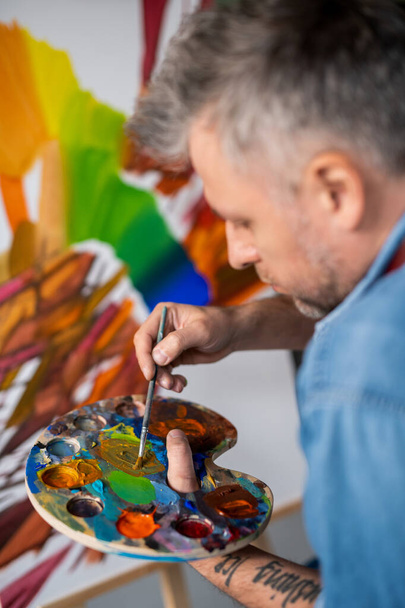 zeitgenössischer professioneller Maler mischt Gouache oder Ölfarbe auf Palette, während er im Atelier ein neues Meisterwerk schafft - Foto, Bild