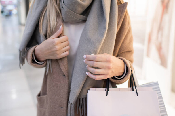 Metà sezione di giovane donna alla moda in cappotto e sciarpa che trasporta sacchetti di carta mentre si muove all'interno del centro commerciale dopo lo shopping
 - Foto, immagini