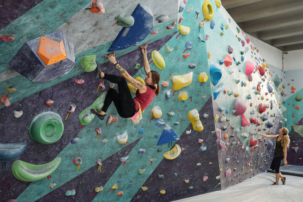 Uma das jovens mulheres ativas praticando escalada no fundo de outra fêmea em pé no tapete na frente de equipamentos esportivos
 - Foto, Imagem