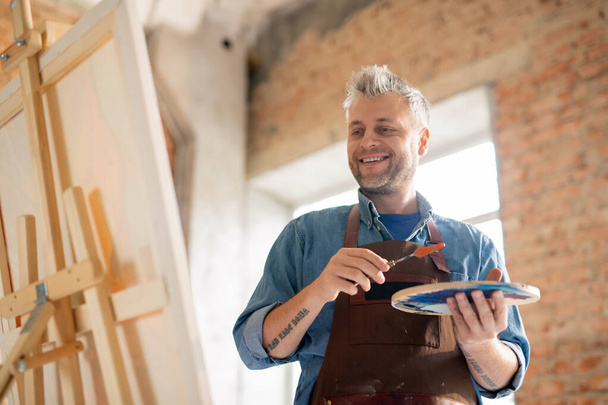 Pintor feliz e bem sucedido com paleta olhando para cavalete enquanto está na frente da pintura em estúdio
 - Foto, Imagem