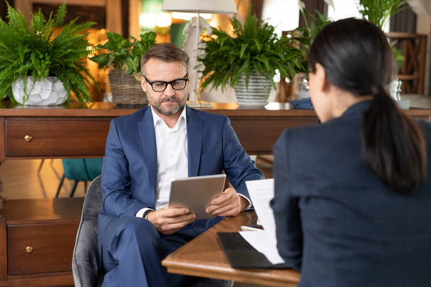 Серьезный бизнесмен среднего возраста или предприниматель с сенсорной панелью просматривает онлайн-информацию с занятым коллегой перед
 - Фото, изображение