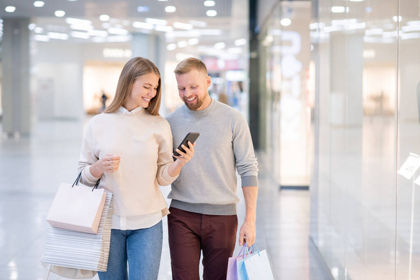 Radosna dziewczyna ze smartfonem i jej chłopakiem przeglądająca towary online w sklepie internetowym podczas relaksu w centrum handlowym - Zdjęcie, obraz