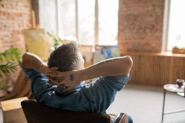 Вид сзади на уставшего художника с грязными руками, сидящего в студии посреди рабочего дня и наслаждающегося отдыхом
 - Фото, изображение