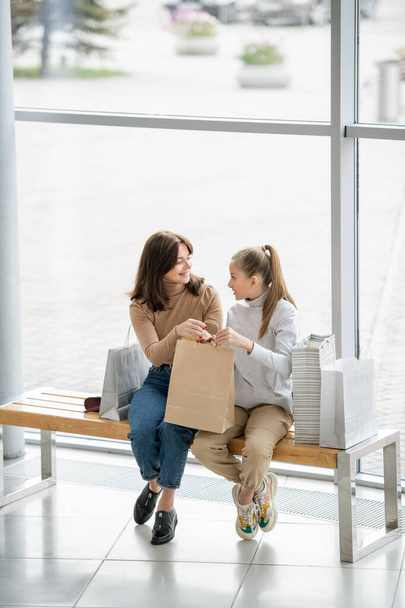 Ładna dziewczyna i jej matka trzymają teczkę, siedząc na ławce i dyskutując o tym, co kupili w centrum handlowym - Zdjęcie, obraz