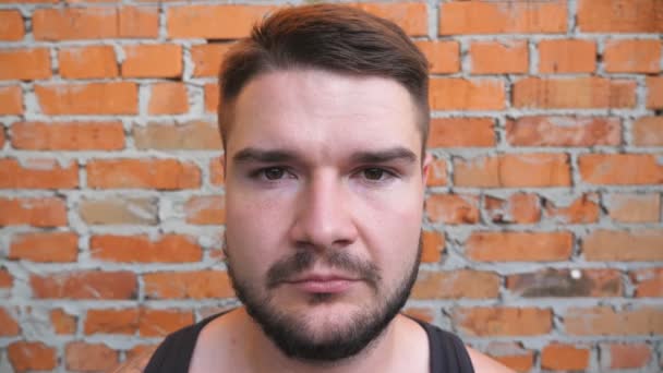 Portrait of young bearded guy looking into camera with confident and serious expression on his face outdoor. Close up of strong man with brown hair standing on blurred brick wall background - Footage, Video