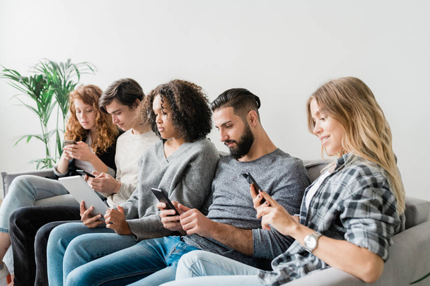 Reihe von fünf lässigen interkulturellen freundlichen Millennials, die in ihren mobilen Geräten scrollen, während sie auf der Couch sitzen - Foto, Bild