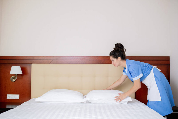 Pretty young woman in uniform of room maid standing by bed and changing pillows in the morning - Photo, Image