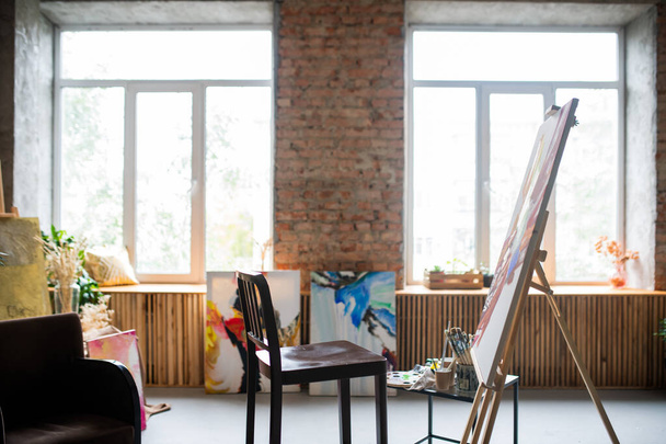Cadeira de madeira do pintor, cavalete com pintura inacabada, vários equipamentos para a arte que trabalha dentro do estúdio ou da oficina
 - Foto, Imagem