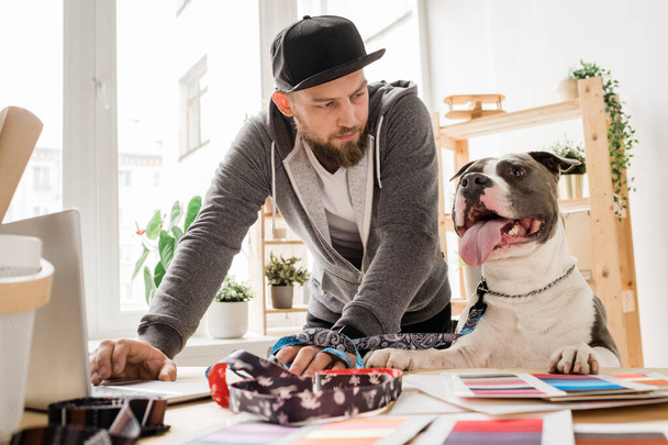 Young casual man looking at his dog while bending over workplace with laptop and working over new collar collection - Фото, изображение