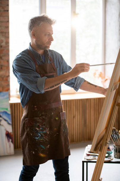Homme d'âge moyen sérieux avec pinceau regardant l'image sur chevalet tout en peignant dans son atelier ou atelier
 - Photo, image