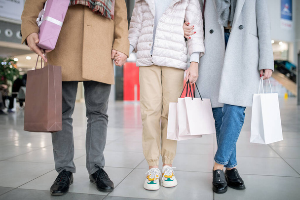 Parte inferior da família casual de três segurando sacos de papel com compras enquanto passa o tempo no shopping depois de fazer compras
 - Foto, Imagem