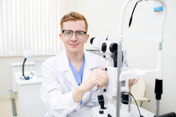 Gelukkig jong stagiaire in whitecoat en bril kijken naar je tijdens het gebruik van nieuwe oftalmologische apparatuur - Foto, afbeelding