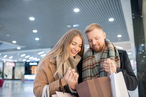 glückliche junge Käufer, die nach dem Einkauf eines der Geschenke in der Papiertüte betrachten, während sie entscheiden, wem sie es schenken können - Foto, Bild
