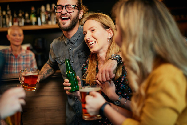 Przystojny biały mężczyzna w okularach przytulający swoją dziewczynę i rozmawiający z nią, stojąc w pubie i pijąc piwo. Nocne życie. - Zdjęcie, obraz
