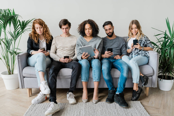 Viisi nuorta nykyajan rento ystäviä istuu sohvalla peräkkäin seinään, kun käytät mobiili vempaimia
 - Valokuva, kuva
