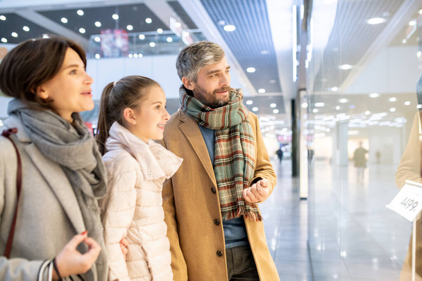 Cheerful family of mother, father and their daughter in casualwear looking at seasonal collection in shop window while chilling in the mall - Фото, изображение