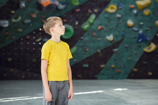 Contemporary active schoolboy in yellow t-shirt and grey pants standing in sports center on background of climbing equipment - Photo, Image